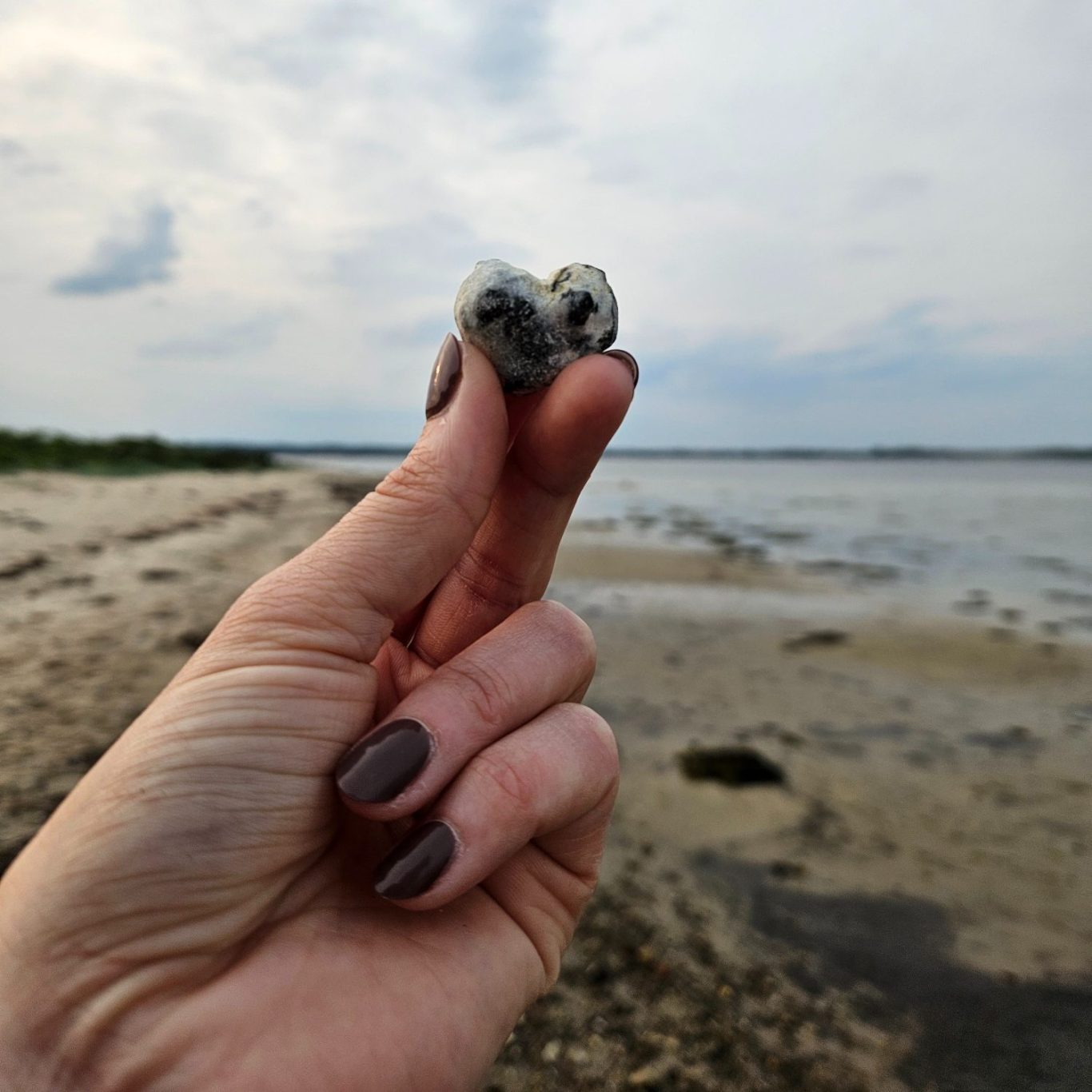 Eine Hand hält einen herzförmigen Stein am Strand, mit Wasser im Hintergrund.
